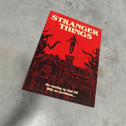 Max Mayfield Running Up That Hill metal poster with a vibrant red Stranger Things design displayed on a textured grey floor.
